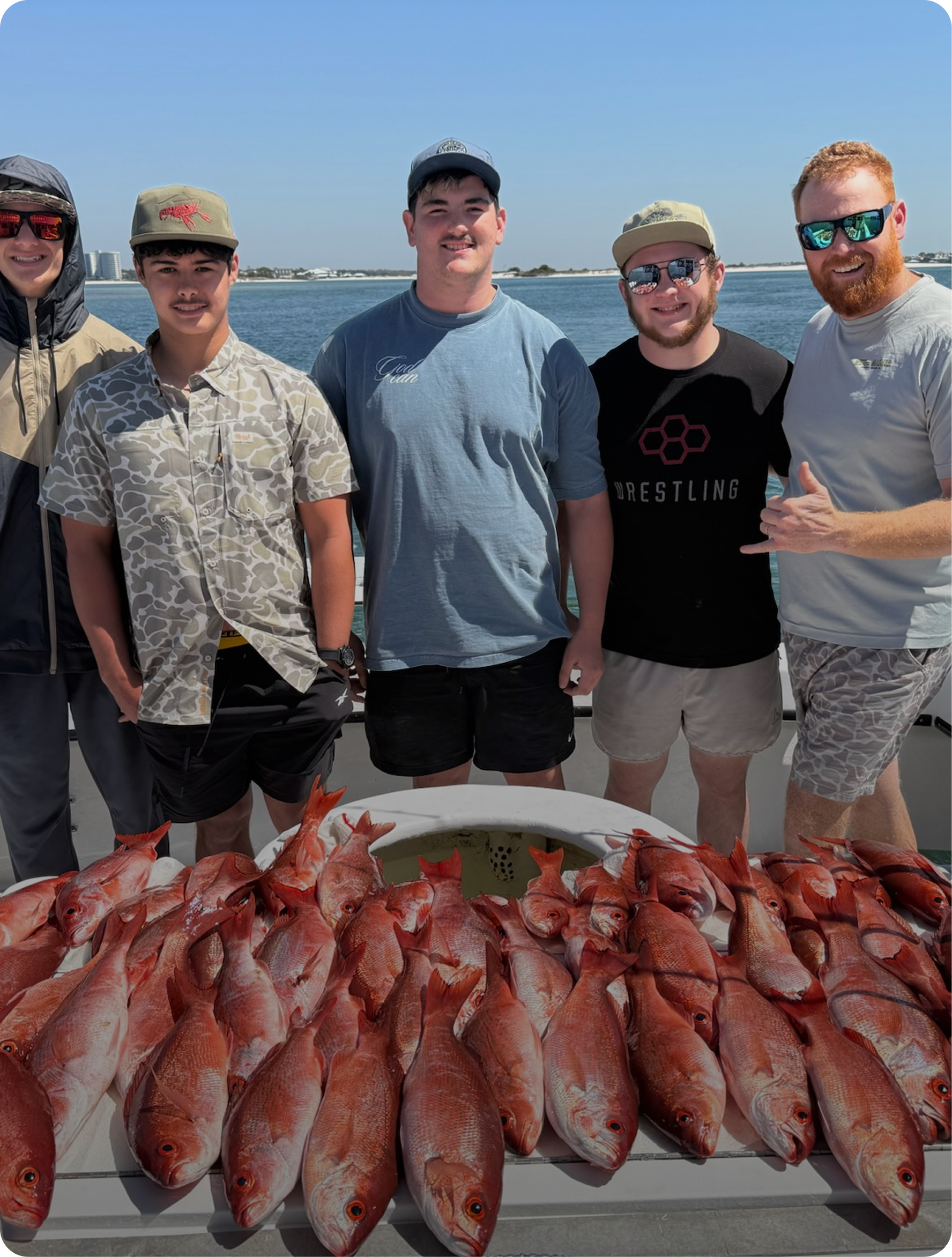 Group of five men standing on a boat with a bounty of red fish laid out on a table in front of them, water and distant shoreline visible behind them on a sunny day.