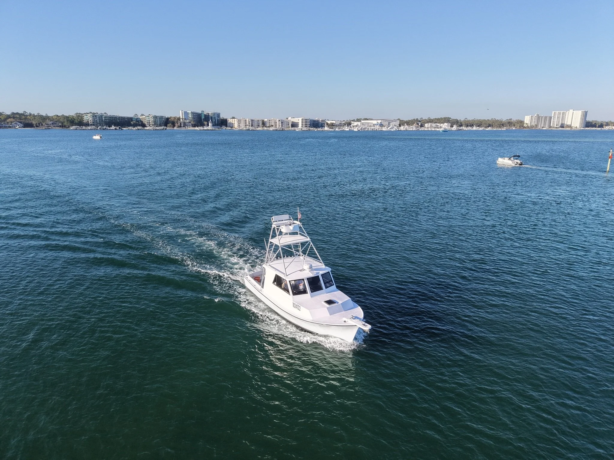 A white boat with a cabin and tower moving through blue water on a sunny day. Several other boats are in the distance, with a shoreline of modern buildings and trees along the horizon.