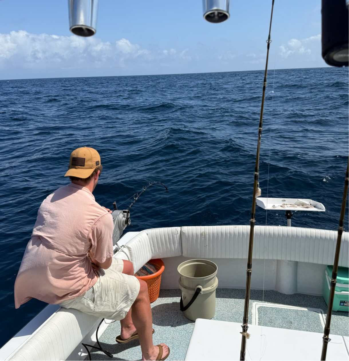 A man in a beige hat and pink shirt fishing from a boat in the open ocean under a partly cloudy sky.