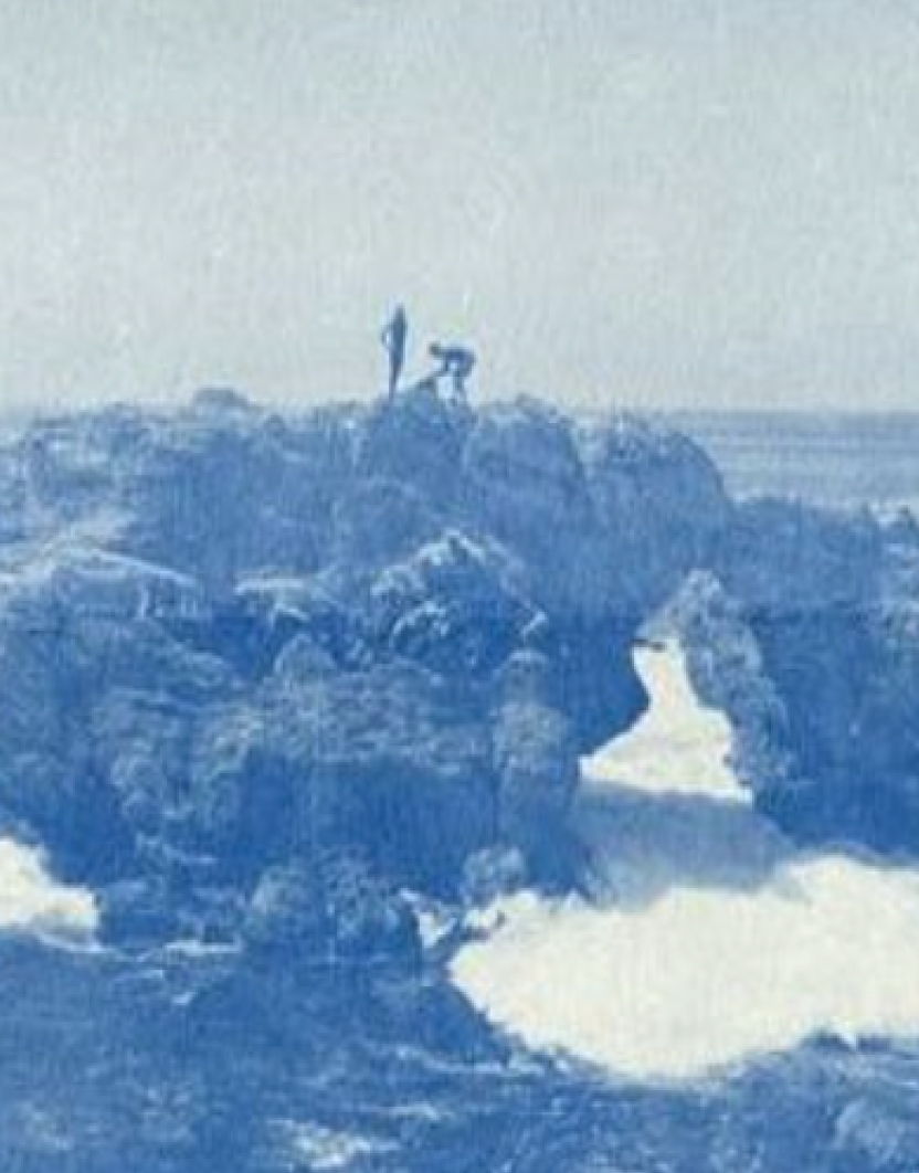 Two people standing and sitting on rocks near the ocean with waves crashing against the rocks, under an overcast sky.
