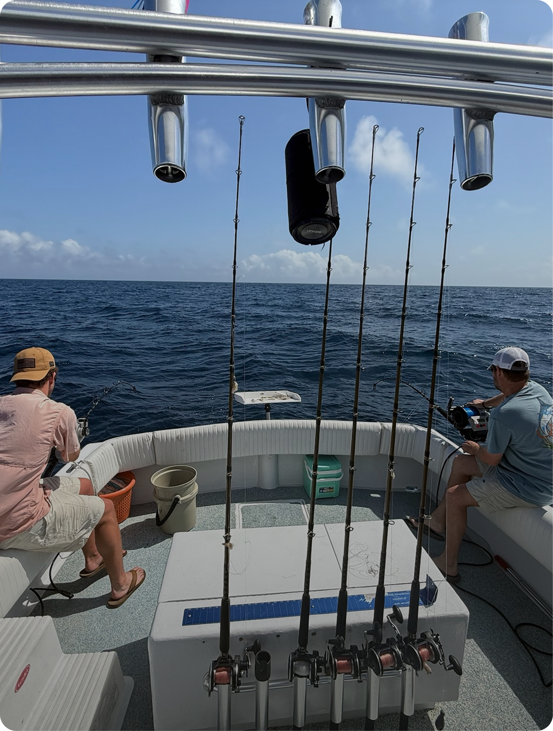Two men fishing on a boat in the open ocean with fishing rods and gear, blue sky, and clouds.