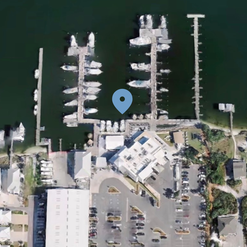 Aerial view of a marina with boats docked along piers, adjacent to a building with a parking lot nearby.