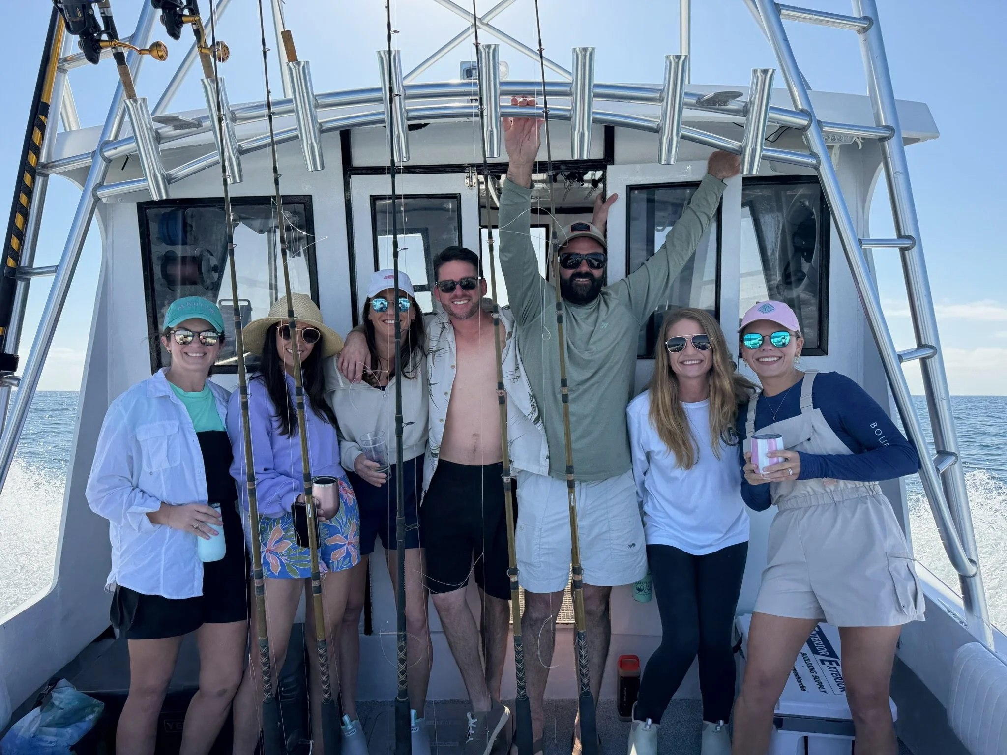 Group of eight friends on a boat with fishing rods, celebrating and smiling at the camera. The sea and sky are visible in the background.