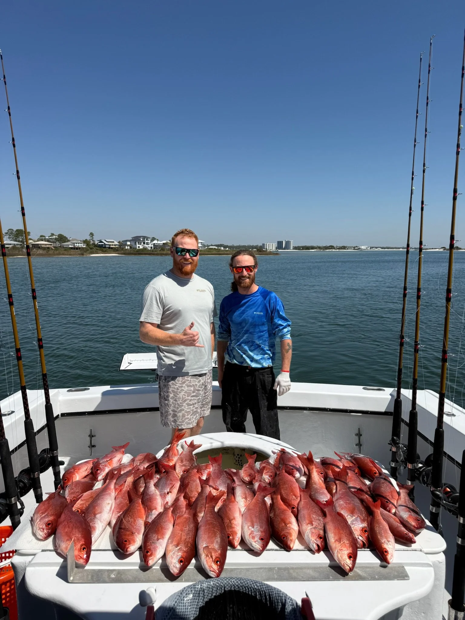 Two men stand on a boat with many red snapper fish laid out on a table, water and shoreline with houses visible in the background, sunny sky.