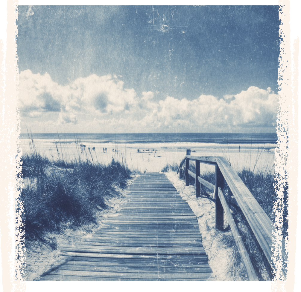 A wooden pathway leading to a beach with sand dunes and grass, with the ocean and sky in the background.