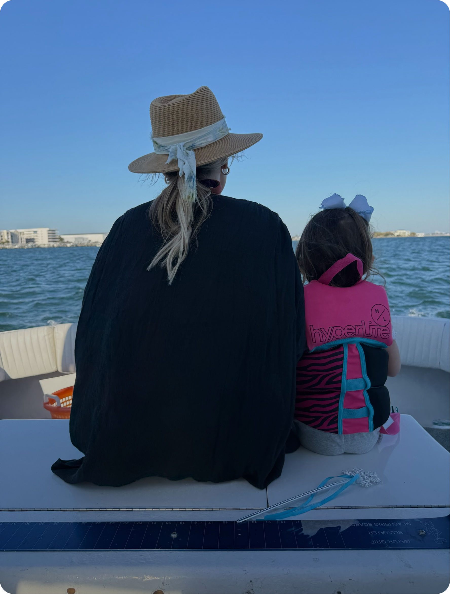 A woman wearing a wide-brimmed straw hat and black jacket sits with a young girl wearing a life jacket, on a boat with a water body and buildings in the background.