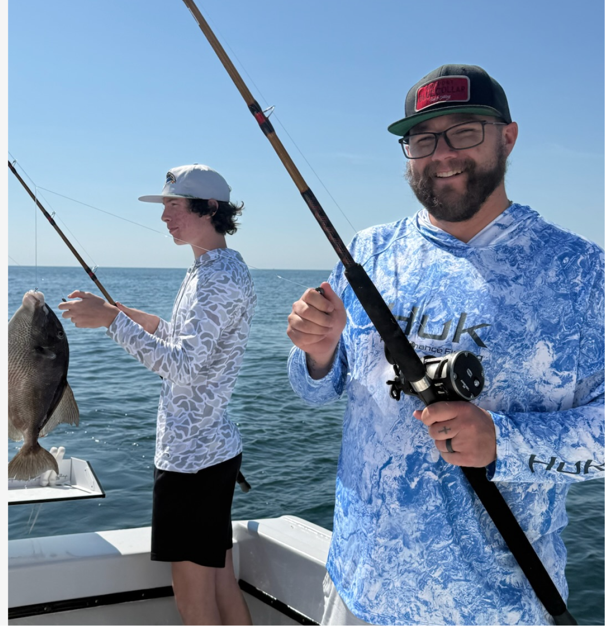 Two men fishing on a boat in the ocean under a clear blue sky, one holding a fishing rod with a fish, and the other looking at his fishing line.