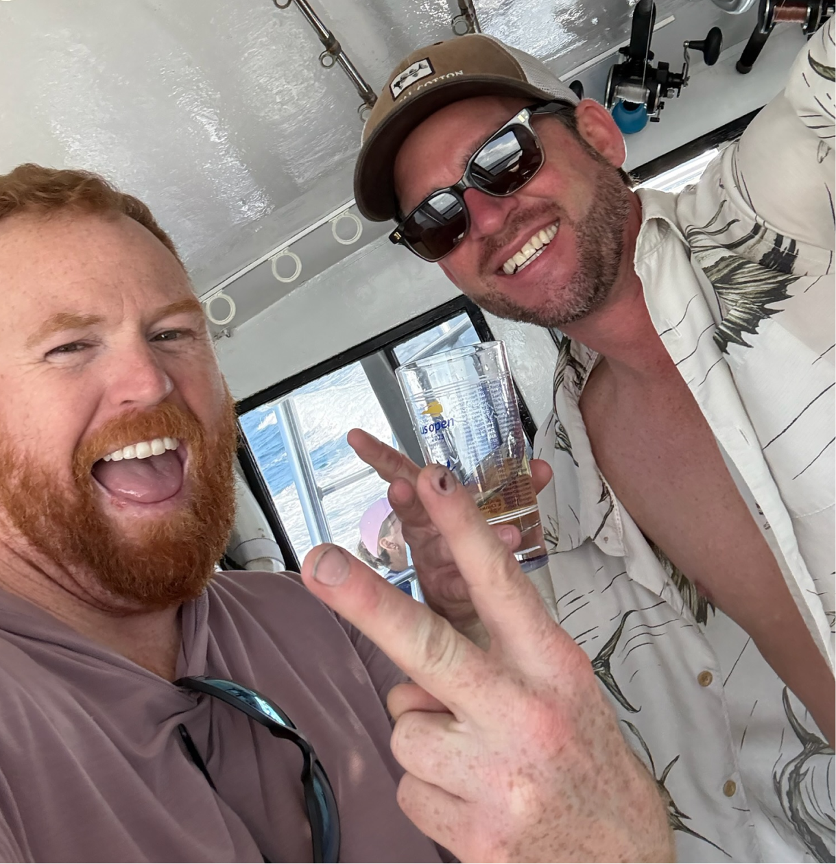 Two men smiling and celebrating on a boat; one with red hair and beard, the other with dark hair, beard, sunglasses, and a cap; one holding a glass of drink in front of the window showing water outside.