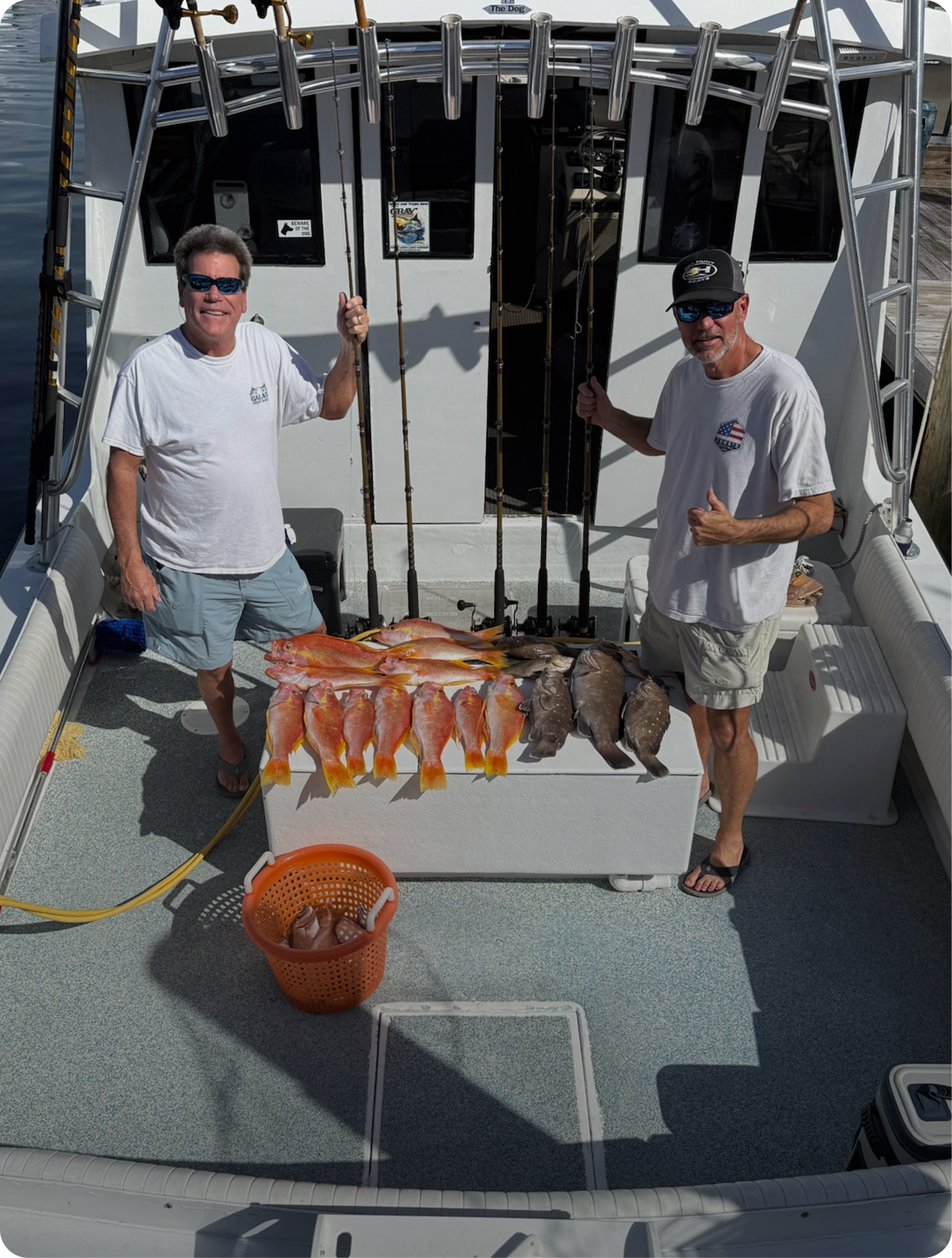 Two men on a boat display their catch of fish, including red and orange fish and darker fish alongside fishing rods.