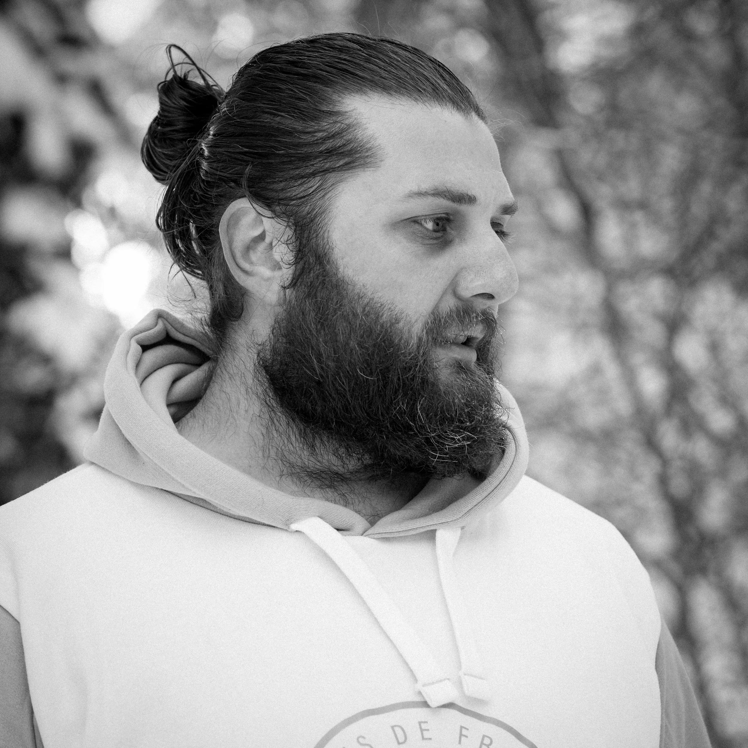 Un homme avec une barbe et des cheveux longs attachés, portant un sweat à capuche, regarde sur le côté, en extérieur.