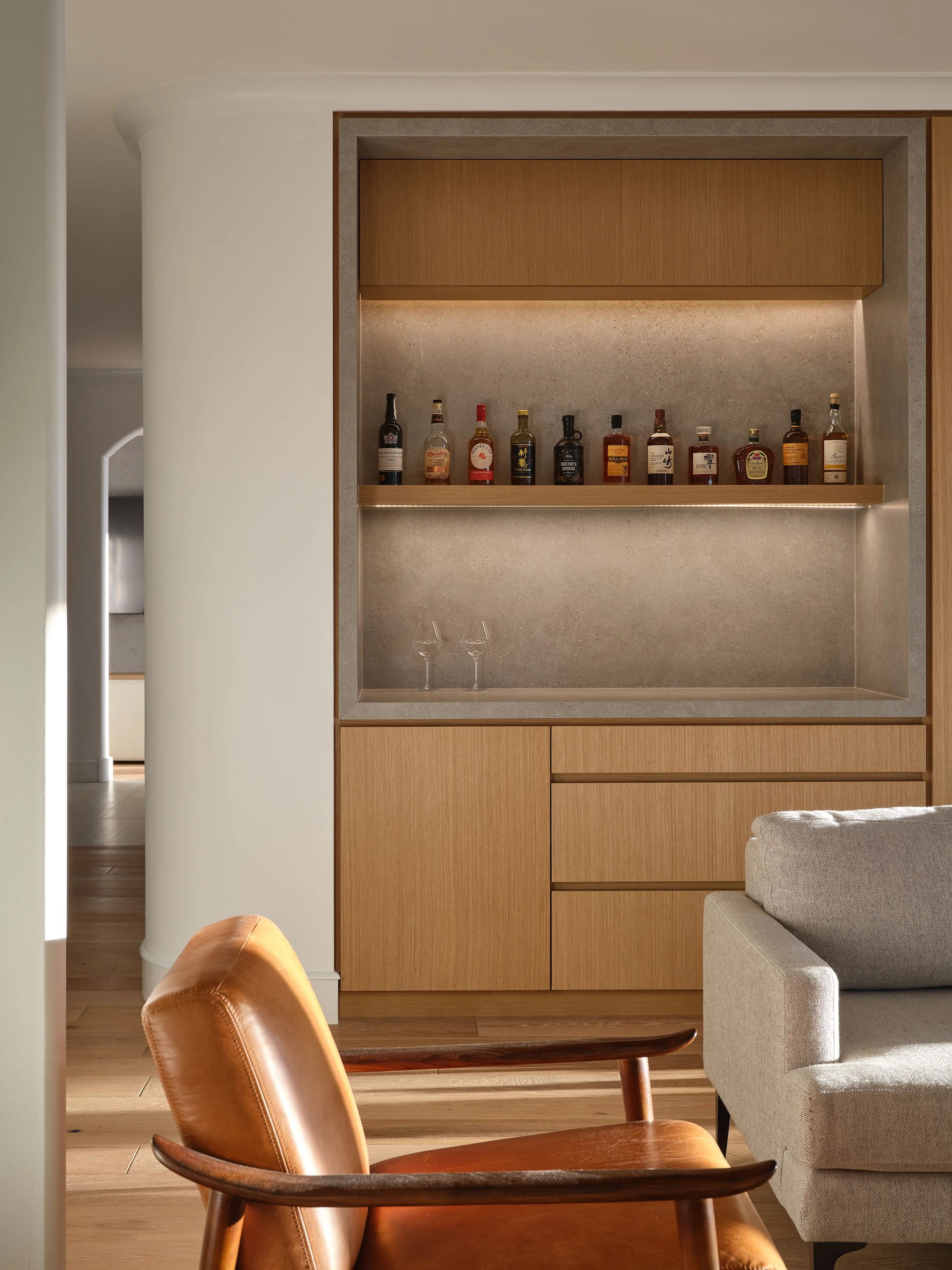 Living room with built-in bar area displaying various liquor bottles on a shelf, with a leather armchair and a beige sofa.