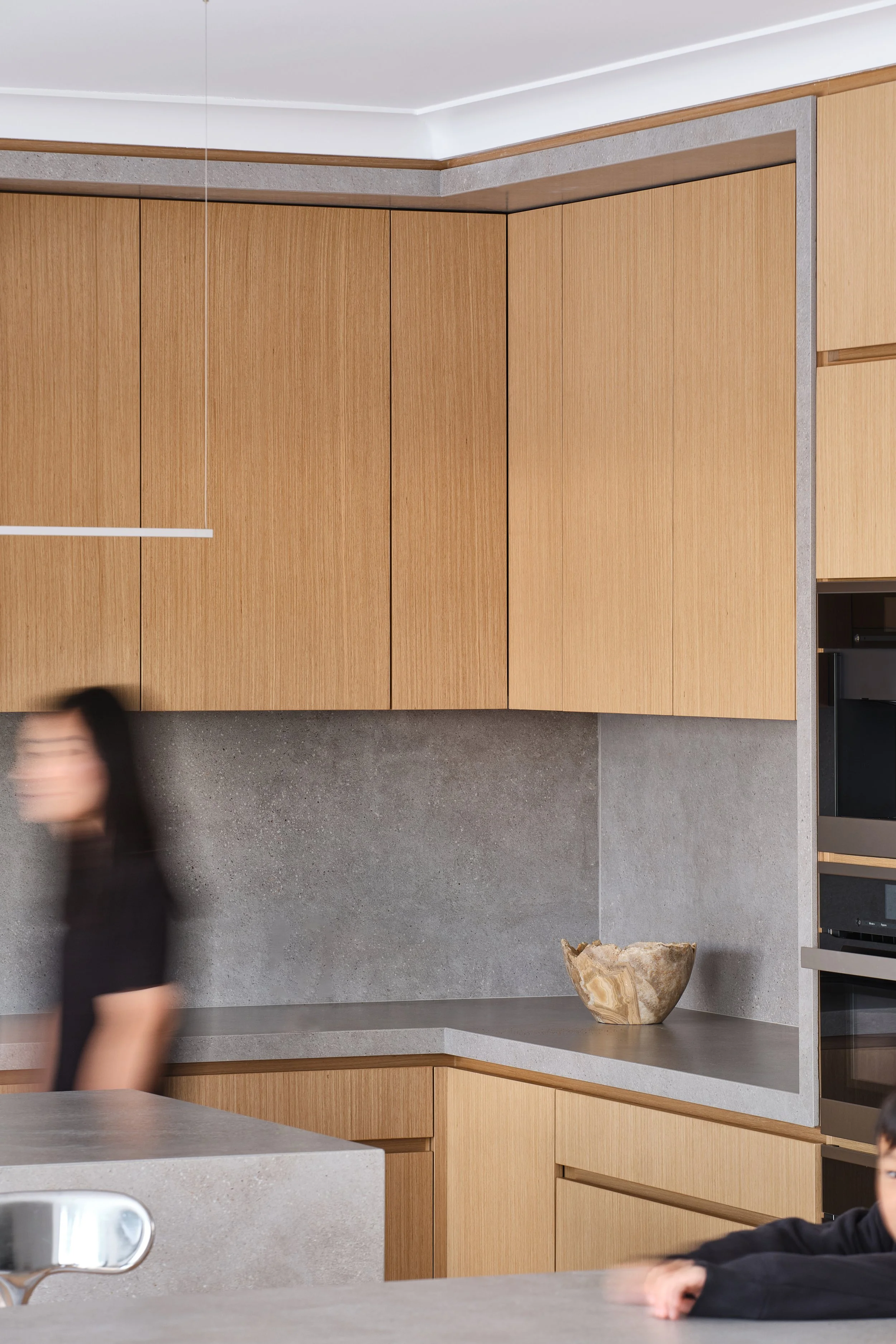 Modern kitchen with wooden cabinets, gray countertops, and a stone backsplash; a blurred woman is walking past and a child is resting on the counter.