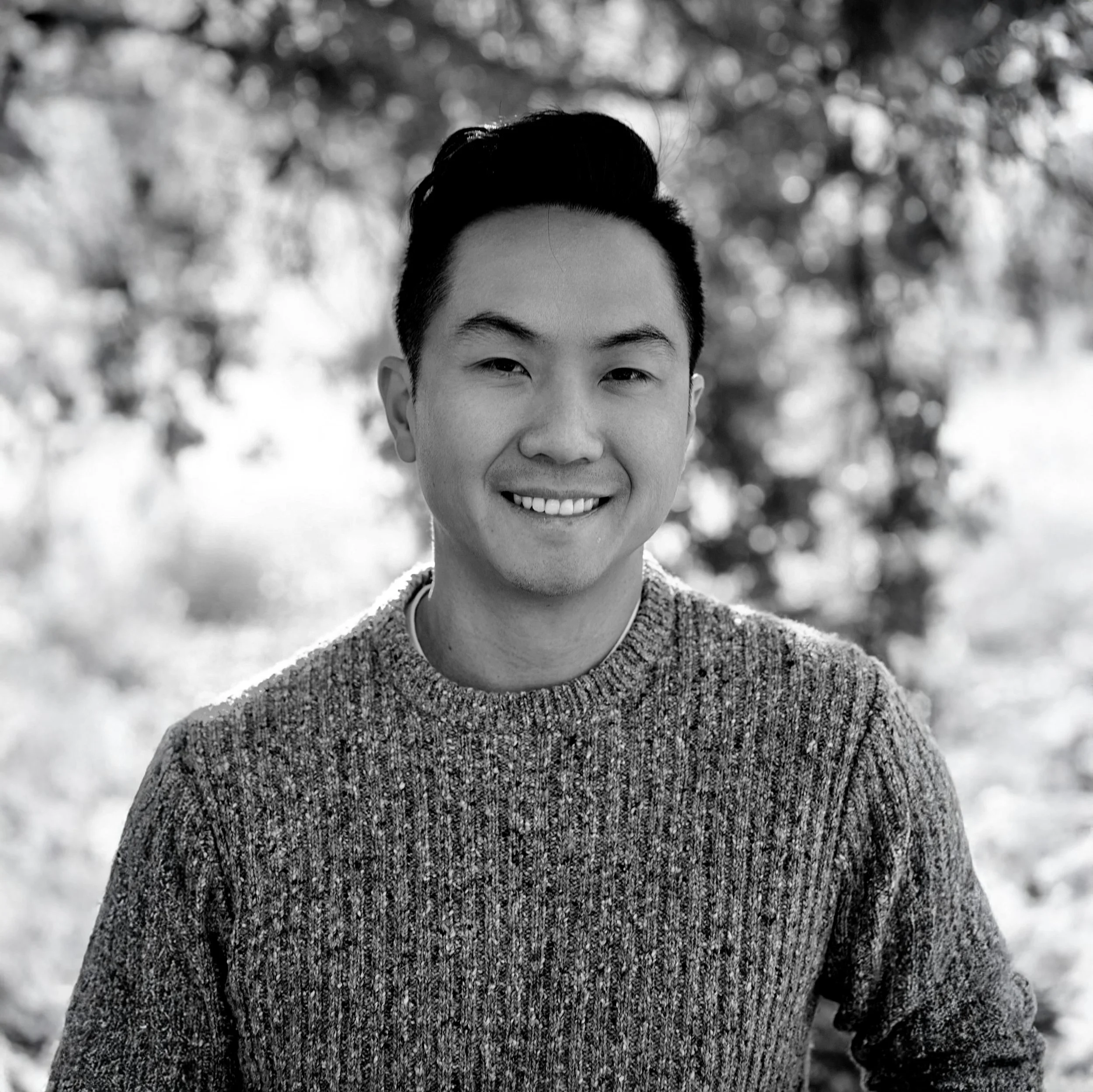 A young man smiling outdoors with trees in the background. He has short, dark hair, is wearing a knit sweater, and is looking at the camera.