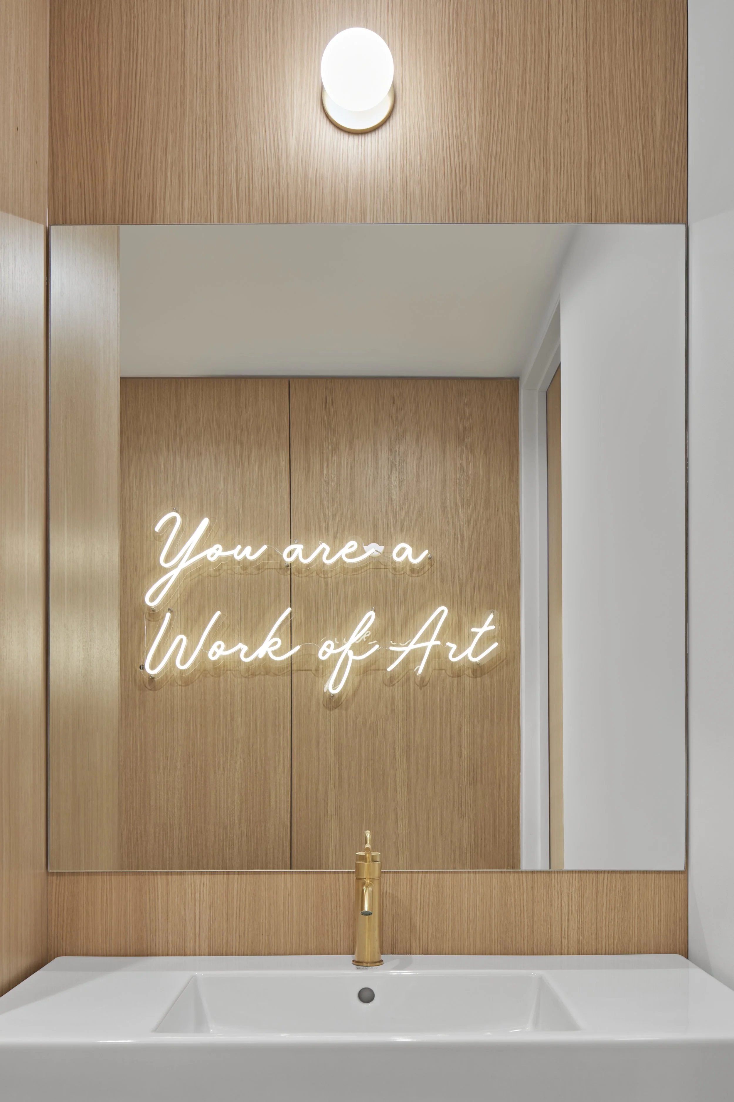 A wall mirror with a neon sign reading "You are a Work of Art." Below the mirror is a white sink with a gold faucet. The wall behind the mirror is made of wood panels, and there is a ceiling light above.