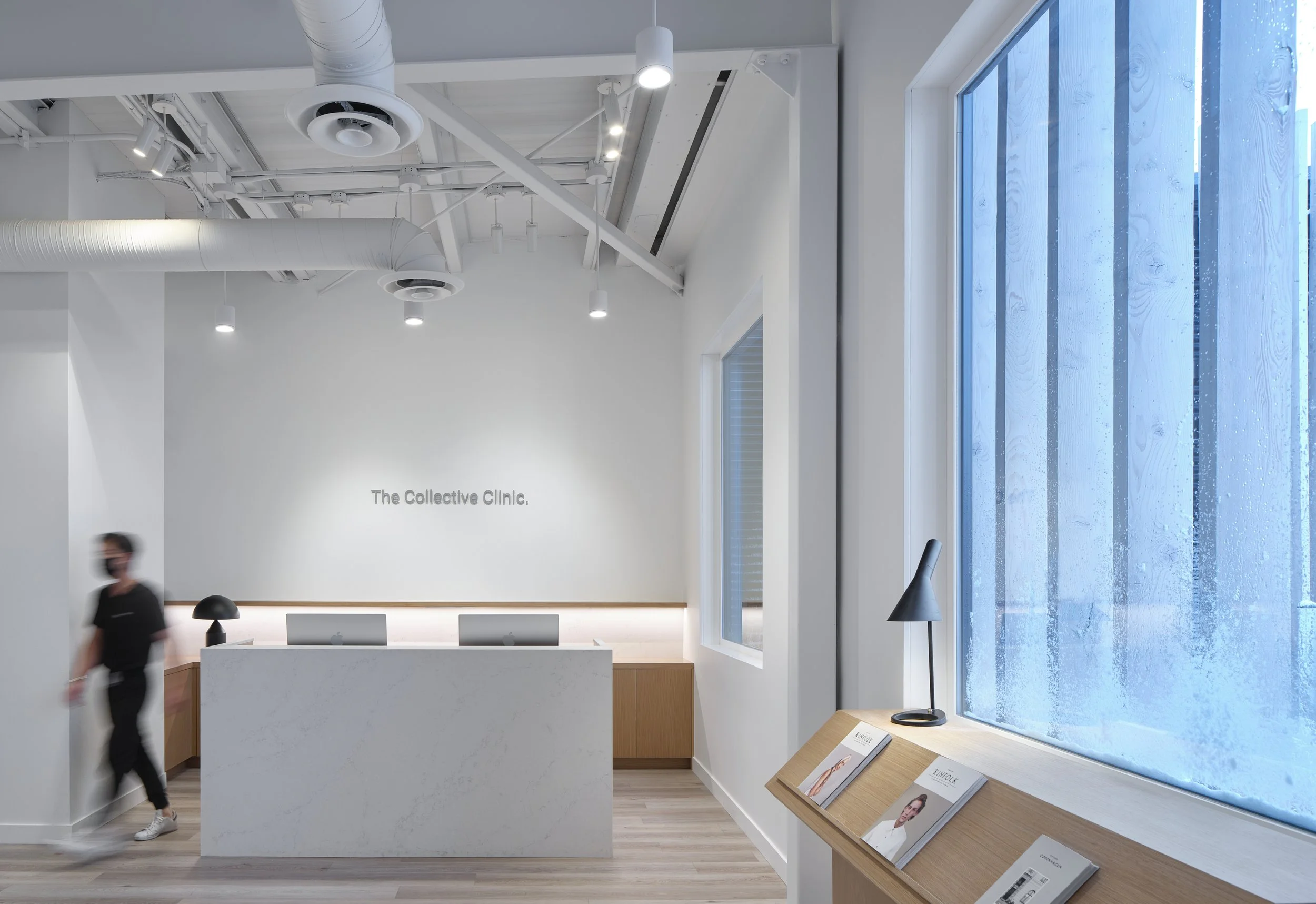 Modern, minimalist clinic reception area with a white desk, two computers, and a person wearing a face mask walking by. The background has a white wall with the text 'The Collective Clinic,' and there is a large window with rain on the glass and a wo