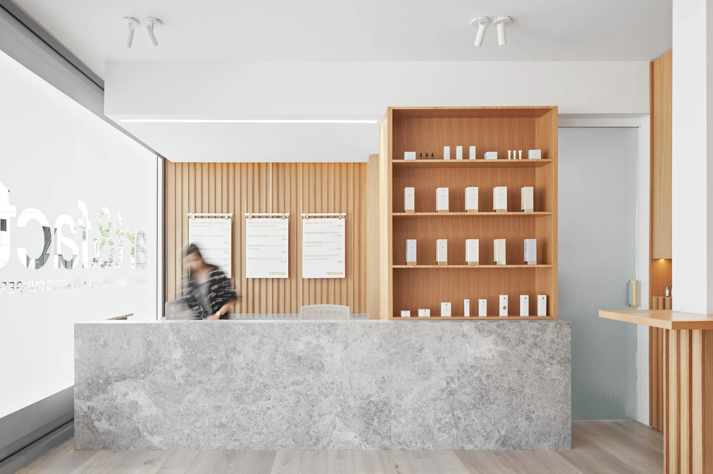 Modern reception area with a marble counter, wooden paneling, and shelves with white product boxes. A blurred person stands behind the counter, and there's a large window to the left with partial text visible.