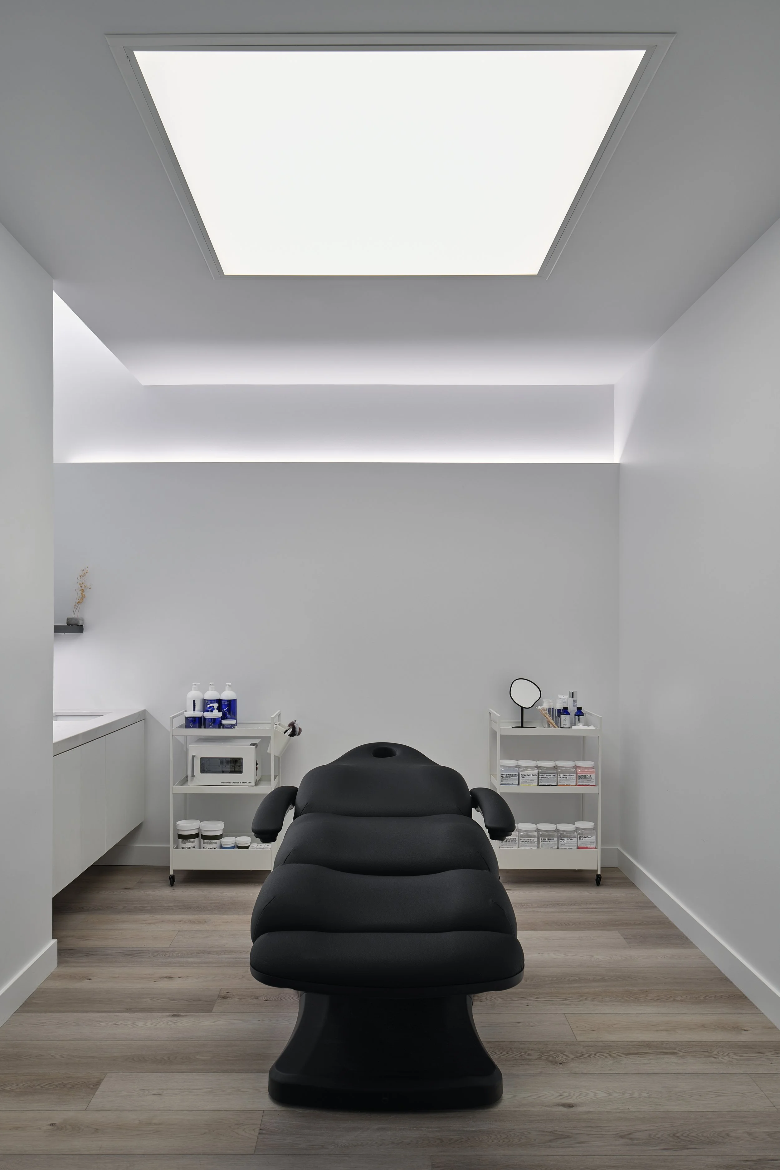 A minimalistic room with a black reclined chair or treatment bed in the center, white walls, and a ceiling with a large light panel. There are white shelves with bottles and jars, a small mirror, and a microwave, suggesting it is a spa or treatment room.