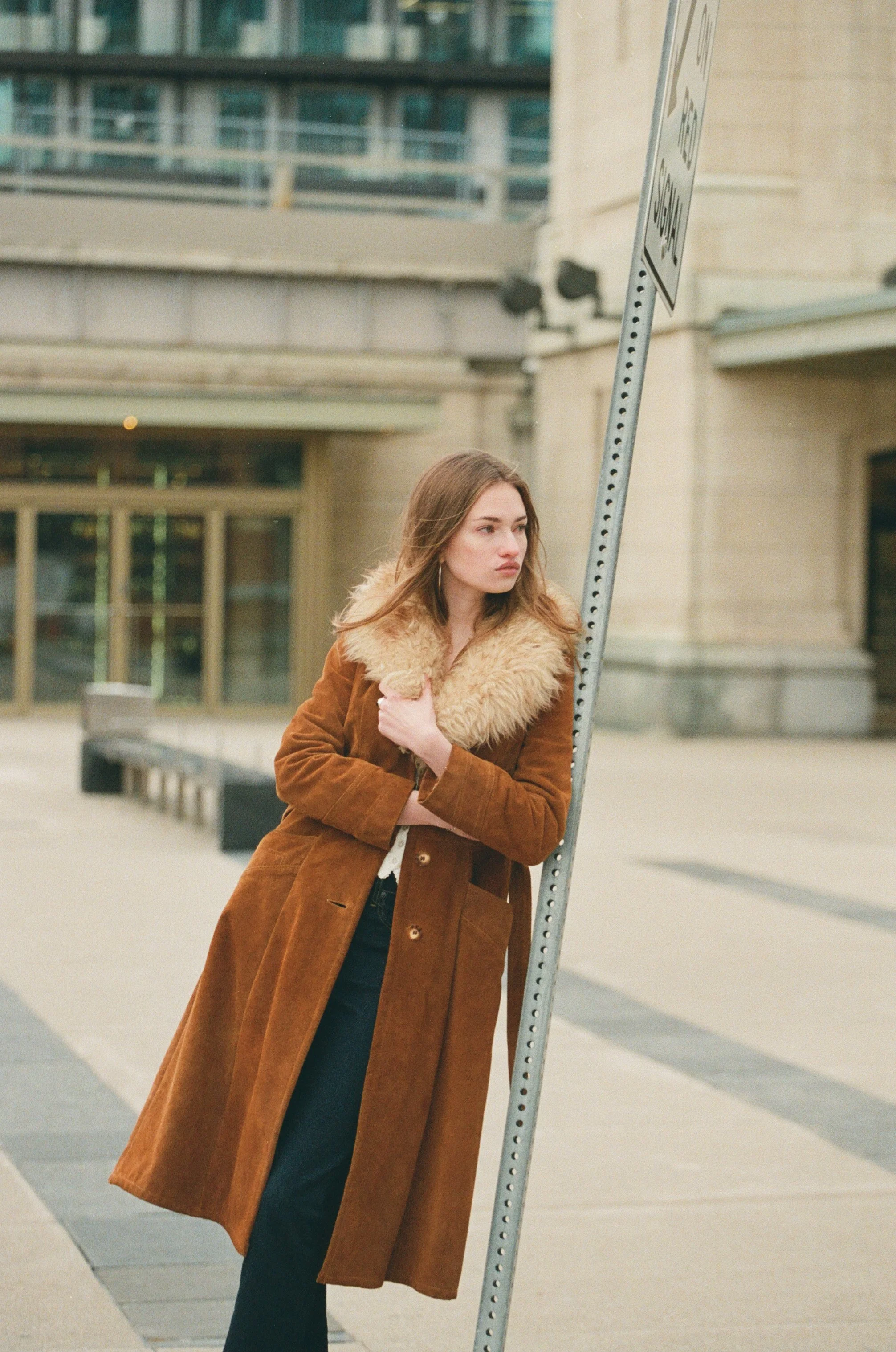 A young woman wearing a brown coat with a fur collar stands next to a street sign on a city sidewalk, with buildings in the background.
