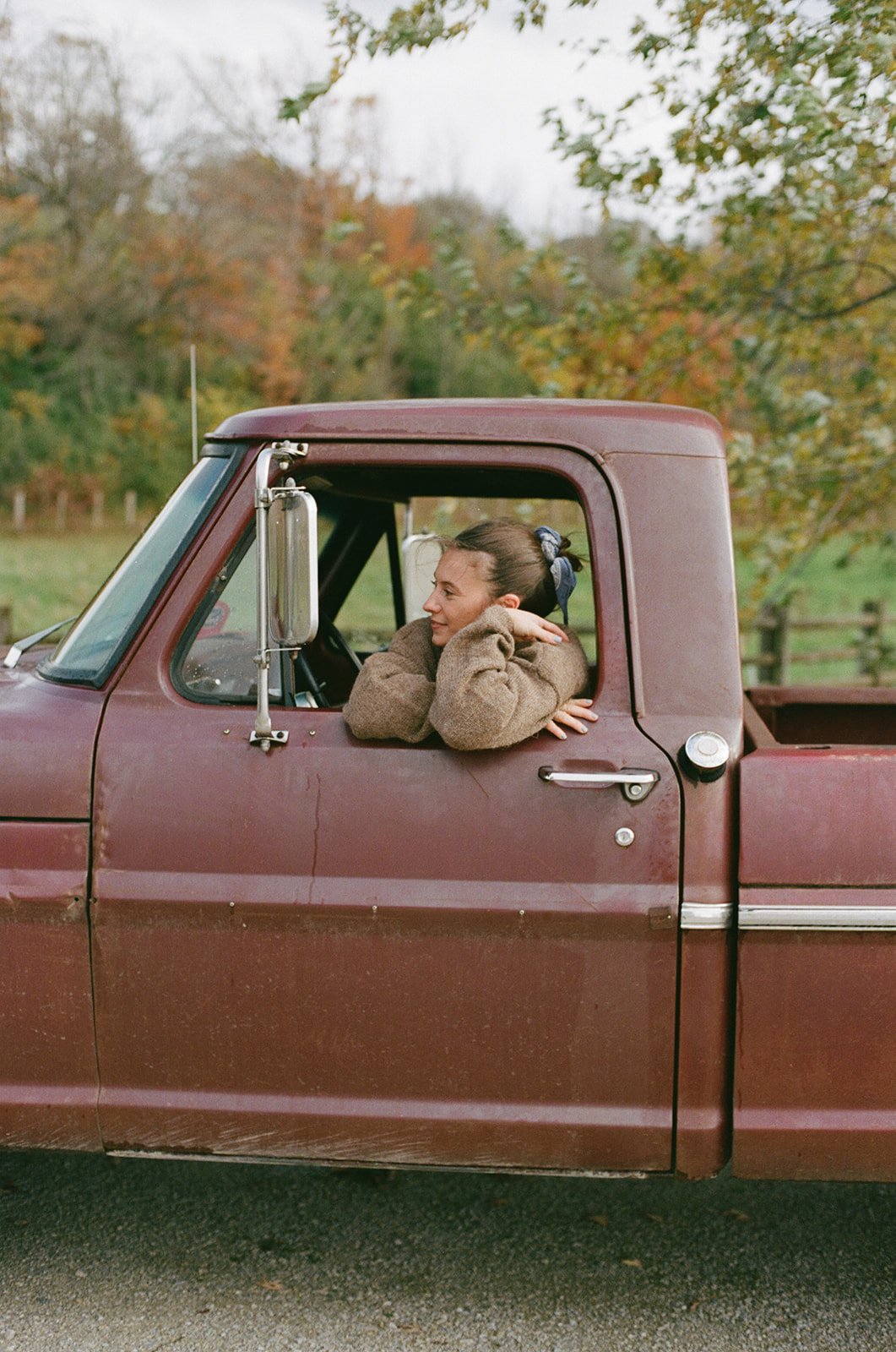 A young woman with a blue scrunchie in her hair leaning on the window of a rusty brown vintage pickup truck, smiling, outdoors in a rural area with trees in autumn colors.