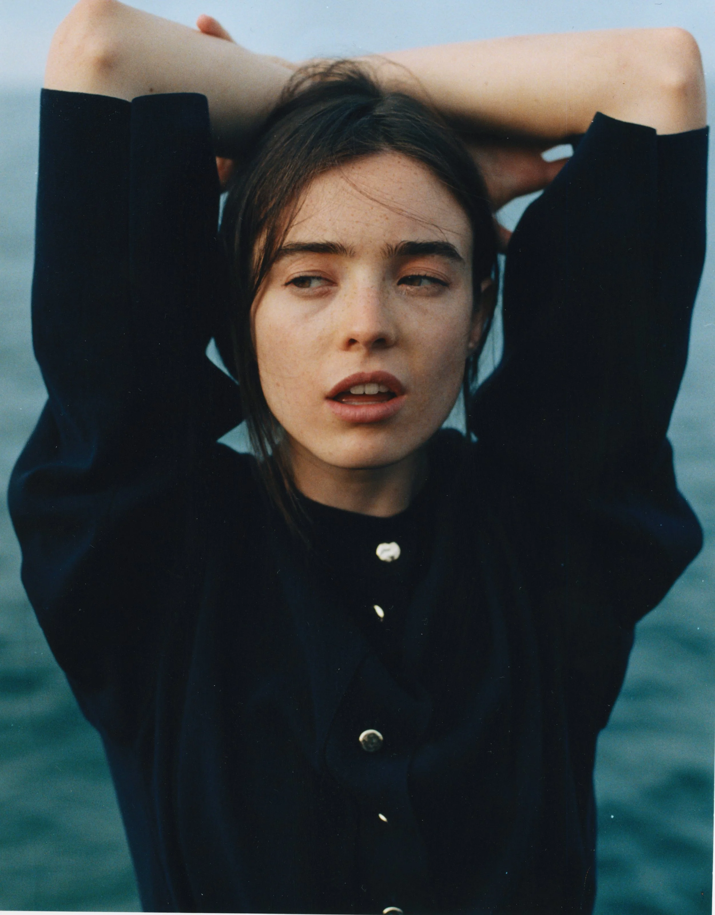 A woman with wet hair and freckles, wearing a black sweater with silver buttons, is standing with her arms raised behind her head against a blue sky and water background.