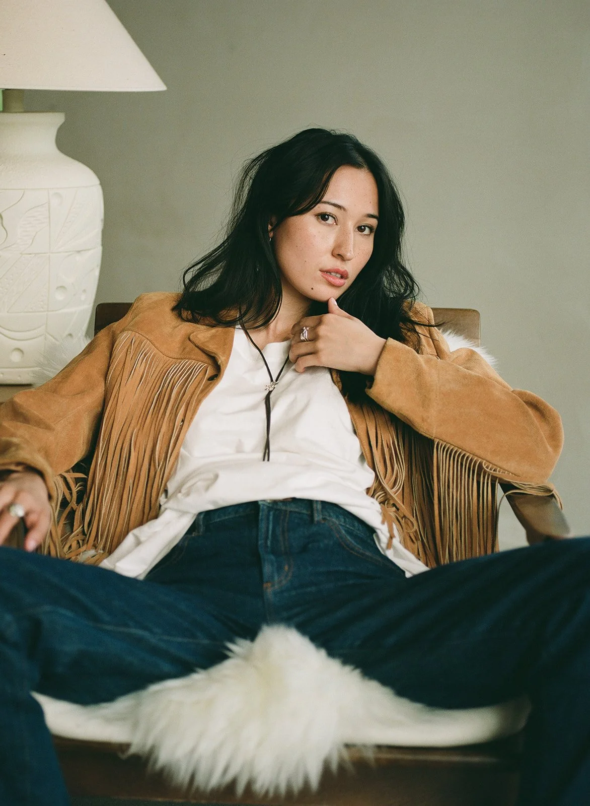 A woman with black hair and light skin sitting on a chair, wearing a tan fringed jacket over a white shirt and blue jeans, with one hand resting on her chest and the other on her lap, in a room with a neutral background and a white lamp.