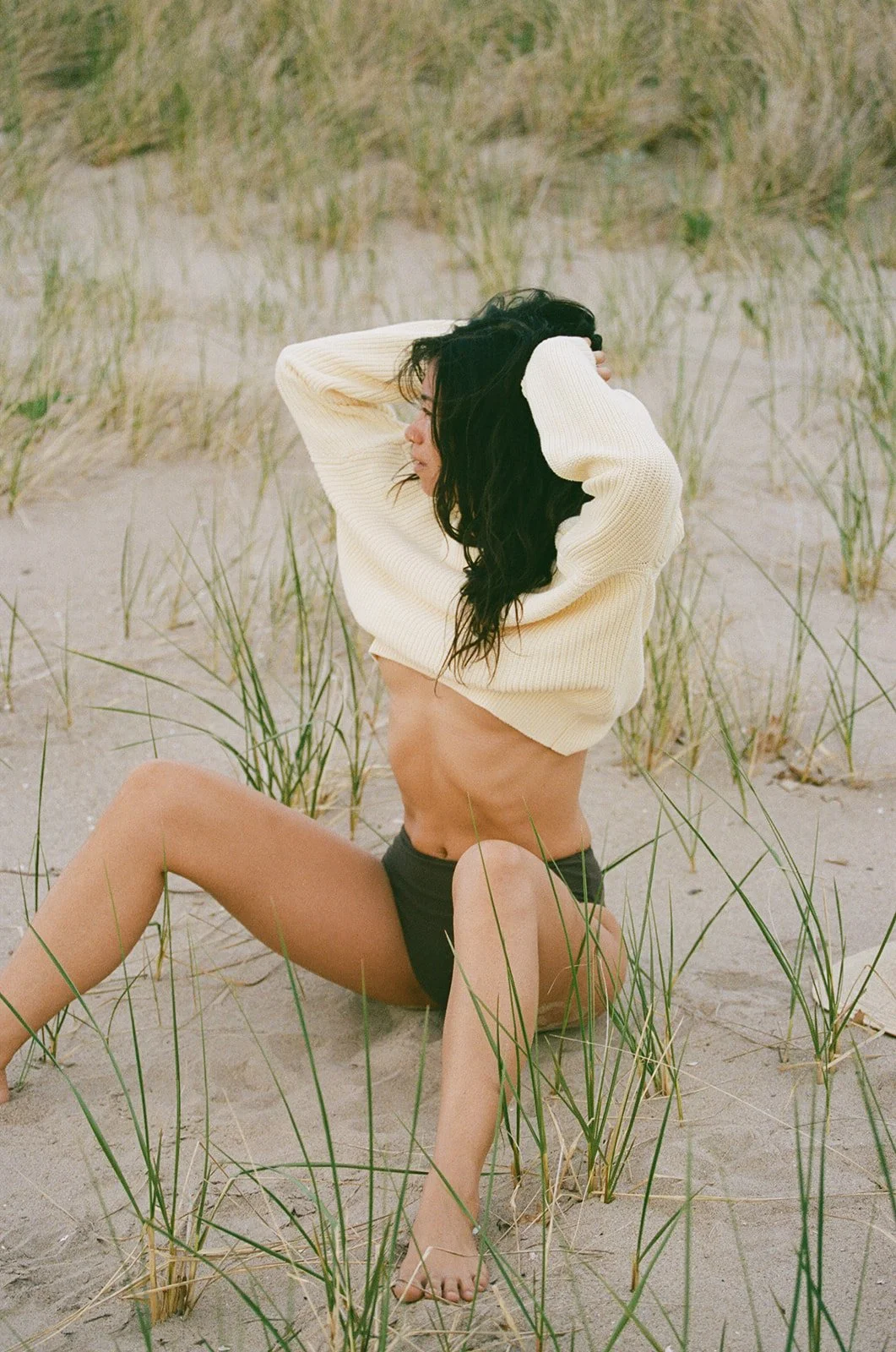 A woman sitting on the sand at a beach with grass in the background, wearing a cream sweater and black shorts, with her hands in her wavy black hair, looking to the side.