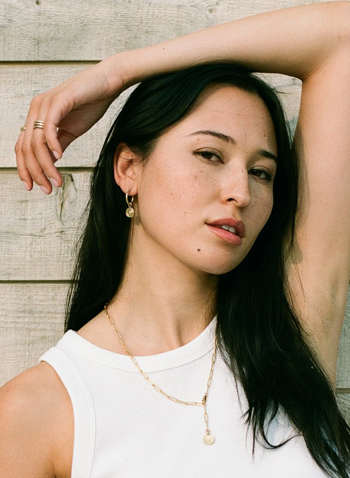 A woman with long dark hair, wearing a white sleeveless top, gold jewelry, and earrings, poses with one arm raised and resting on her head against a wooden background.