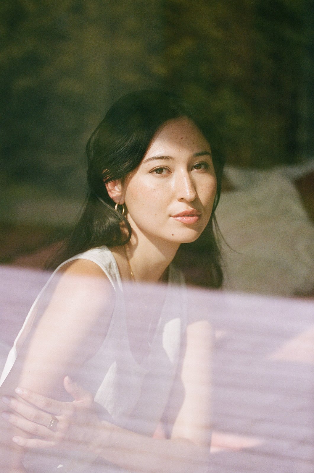 A woman with dark wavy hair, wearing a sleeveless top and gold jewelry, gazes at the camera with a subtle smile, outdoors with a blurred background.