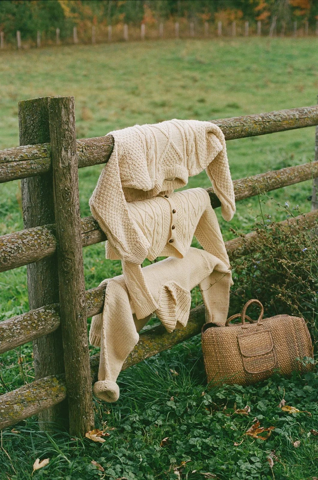 Cream-colored knit sweaters hanging on a wooden fence in a grassy field with oak leaves and a woven bag on the ground.