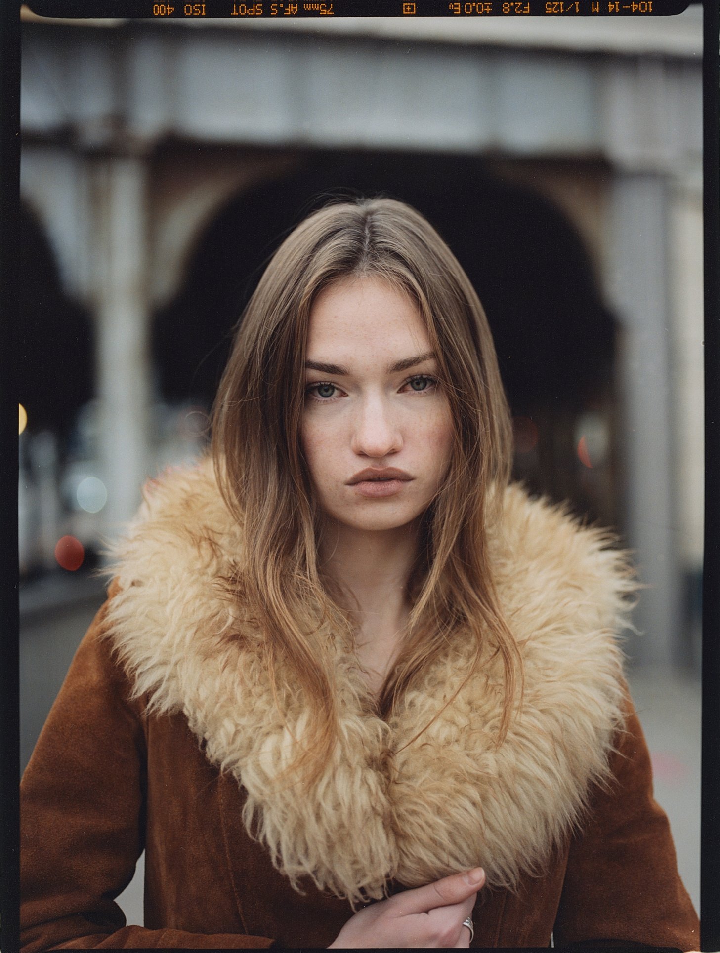 A young woman with long brown hair and light skin is standing outdoors in front of an arched bridge. She is wearing a tan coat with a furry collar. Her expression is neutral, and she is looking directly at the camera.