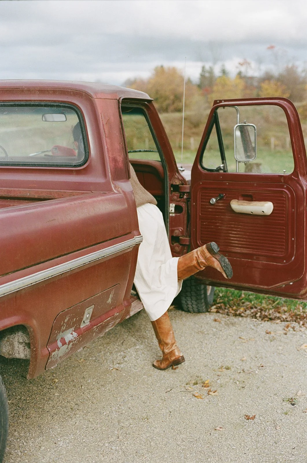 Close-up of an old rusty red pickup truck with a person with one leg sticking out of the open passenger door. The person is wearing tall brown leather boots and white clothing. The background shows a grassy field and trees with autumn foliage under a