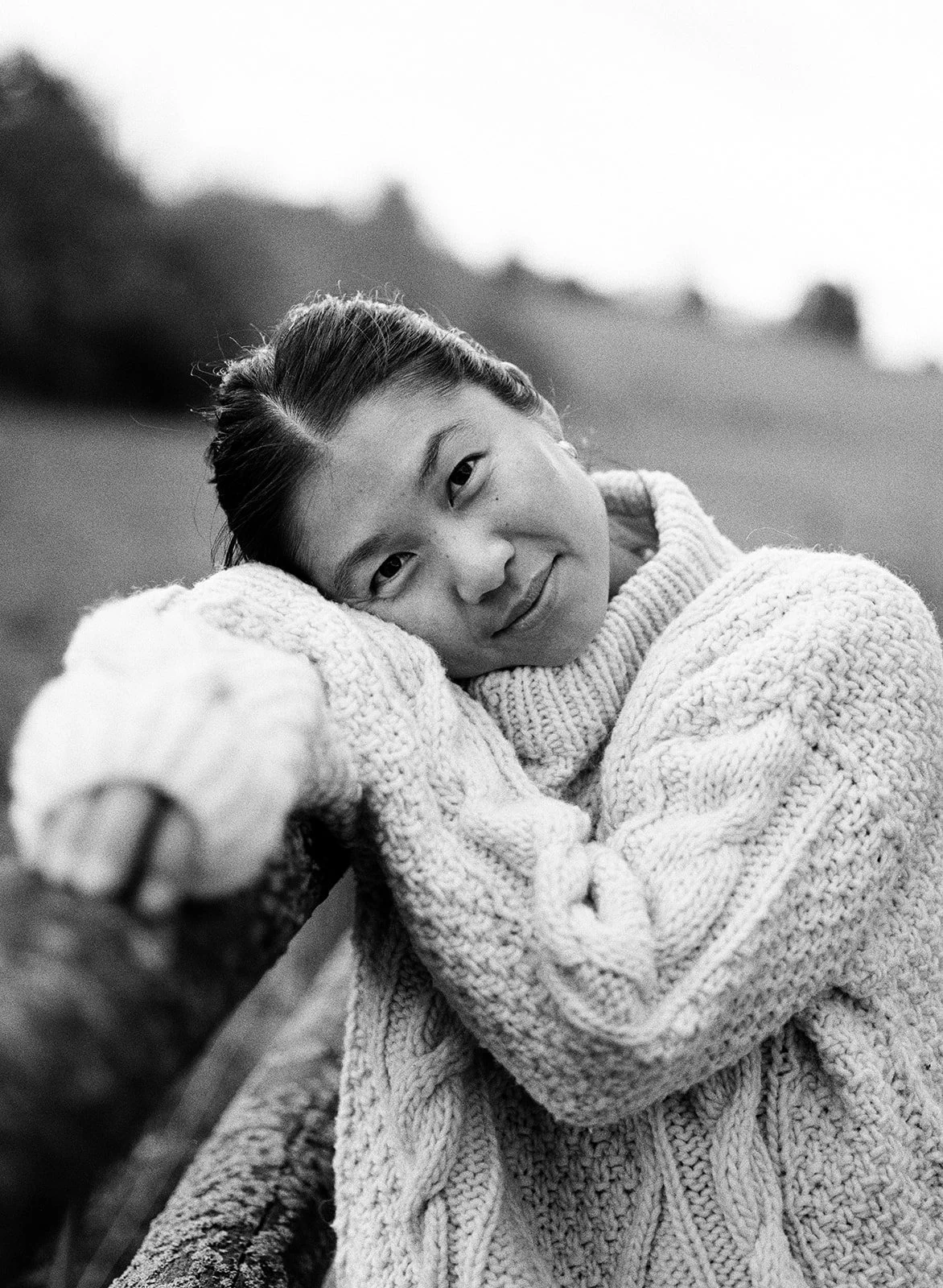 A woman leaning on a log outdoors, wearing a chunky knit sweater, smiling softly at the camera in black and white.