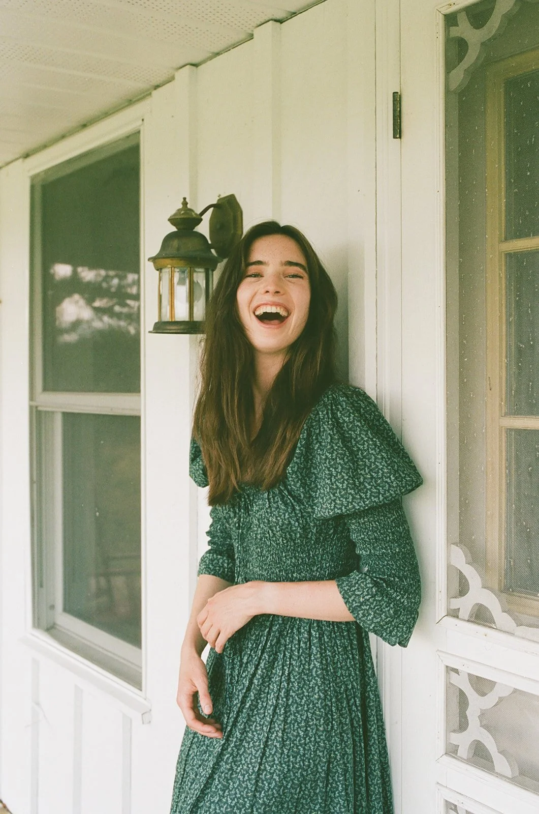 A young woman with long brown hair laughing while standing on a porch, wearing a dark green patterned dress, beside a white wall with a window and an antique lantern.
