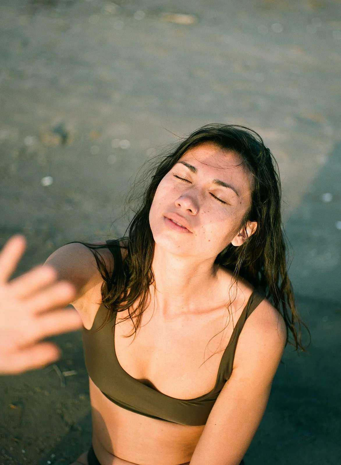 A woman with dark hair and freckles closes her eyes and smiles while taking a selfie on the beach with her arm extended, wearing a brown swimsuit.