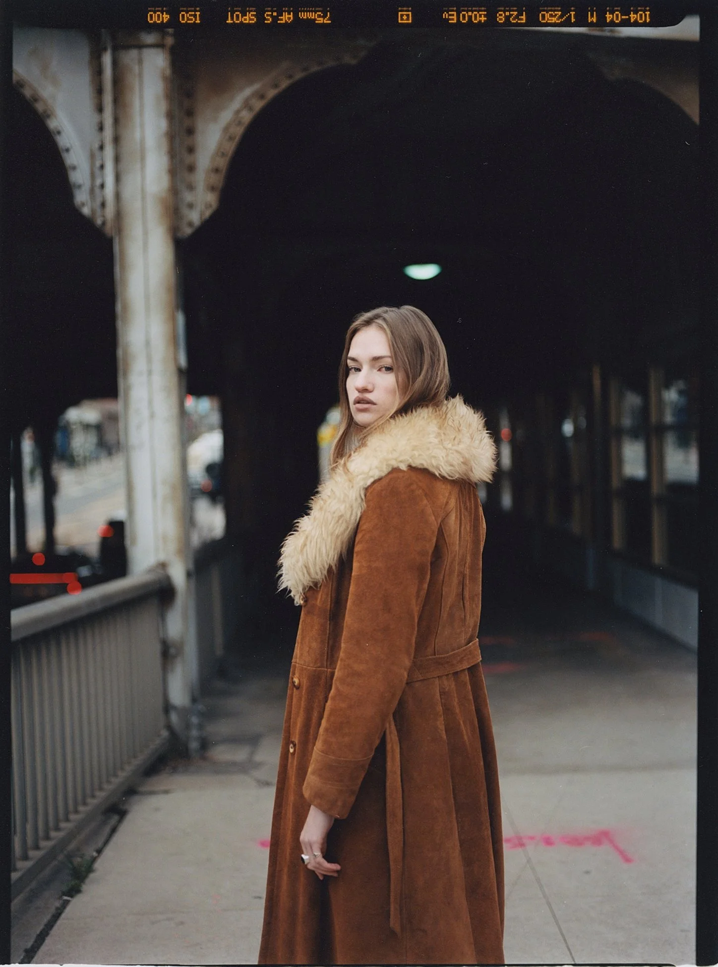 A young woman with fair skin and light brown hair stands on a city street under a bridge, wearing a long brown coat with a faux fur collar. Her expression is serious as she looks toward the camera, with her left hand visible at her side. The backgrou