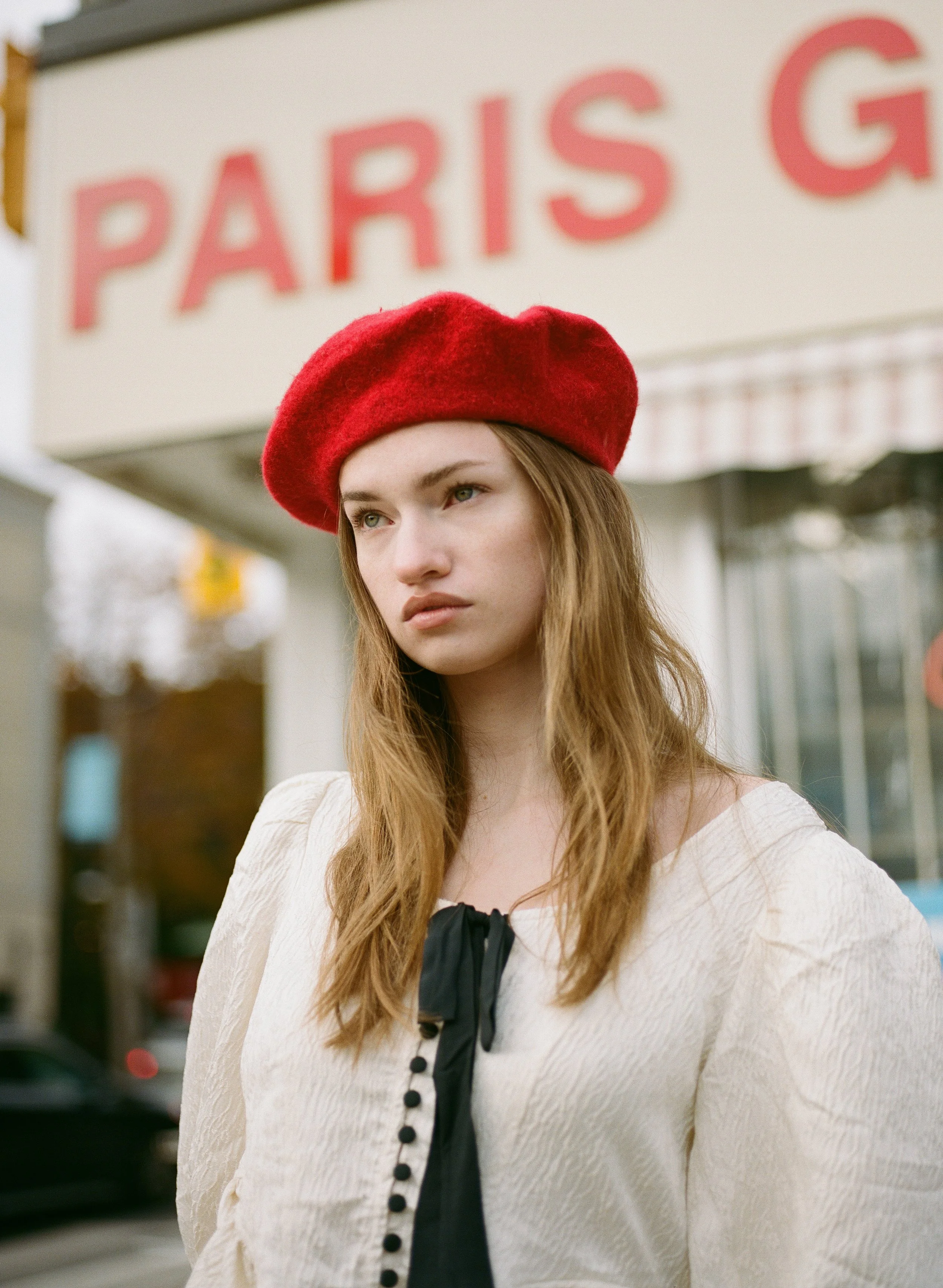 A young woman with long, wavy hair and light skin wearing a red beret and a cream-colored top with black buttons and black ribbon detail, standing outdoors near a Paris sign.