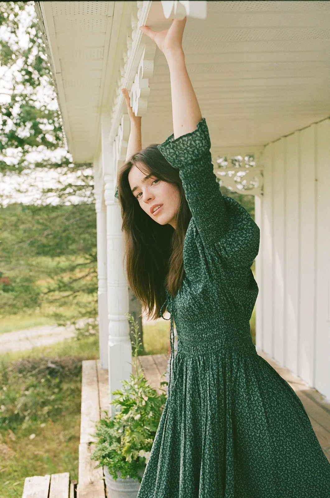 A woman in a green dress with a floral pattern, standing outside on a porch, holding onto the porch railing with one hand while looking at the camera.