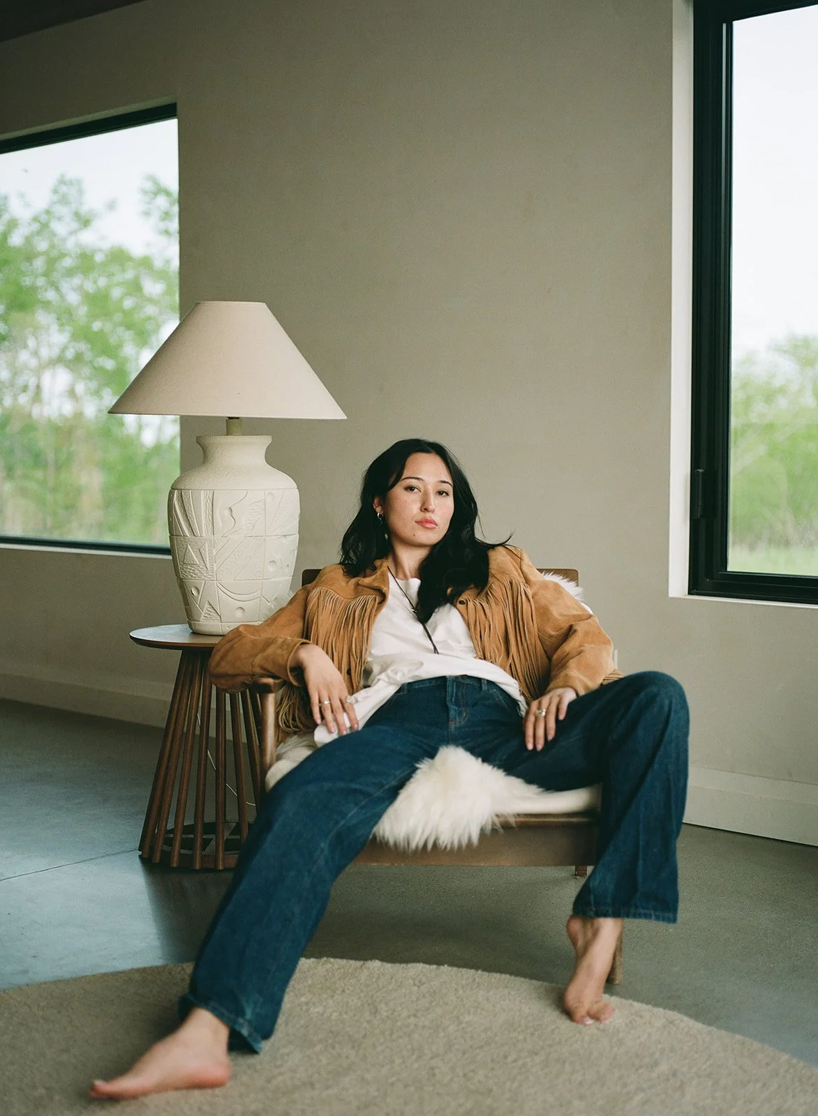 A woman with long black hair dressed in casual clothing, sitting on a chair with bare feet, in a room with large windows and natural light.