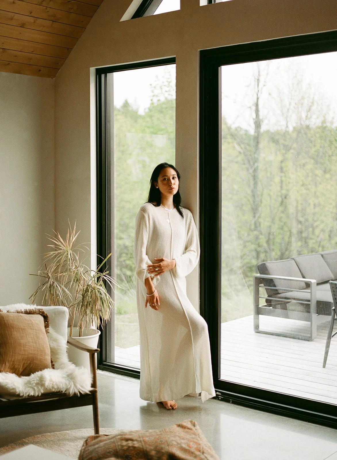 A woman in a cream-colored dress standing near large glass doors, with a mountain of greenery outside, inside a modern home with wooden and neutral decor.