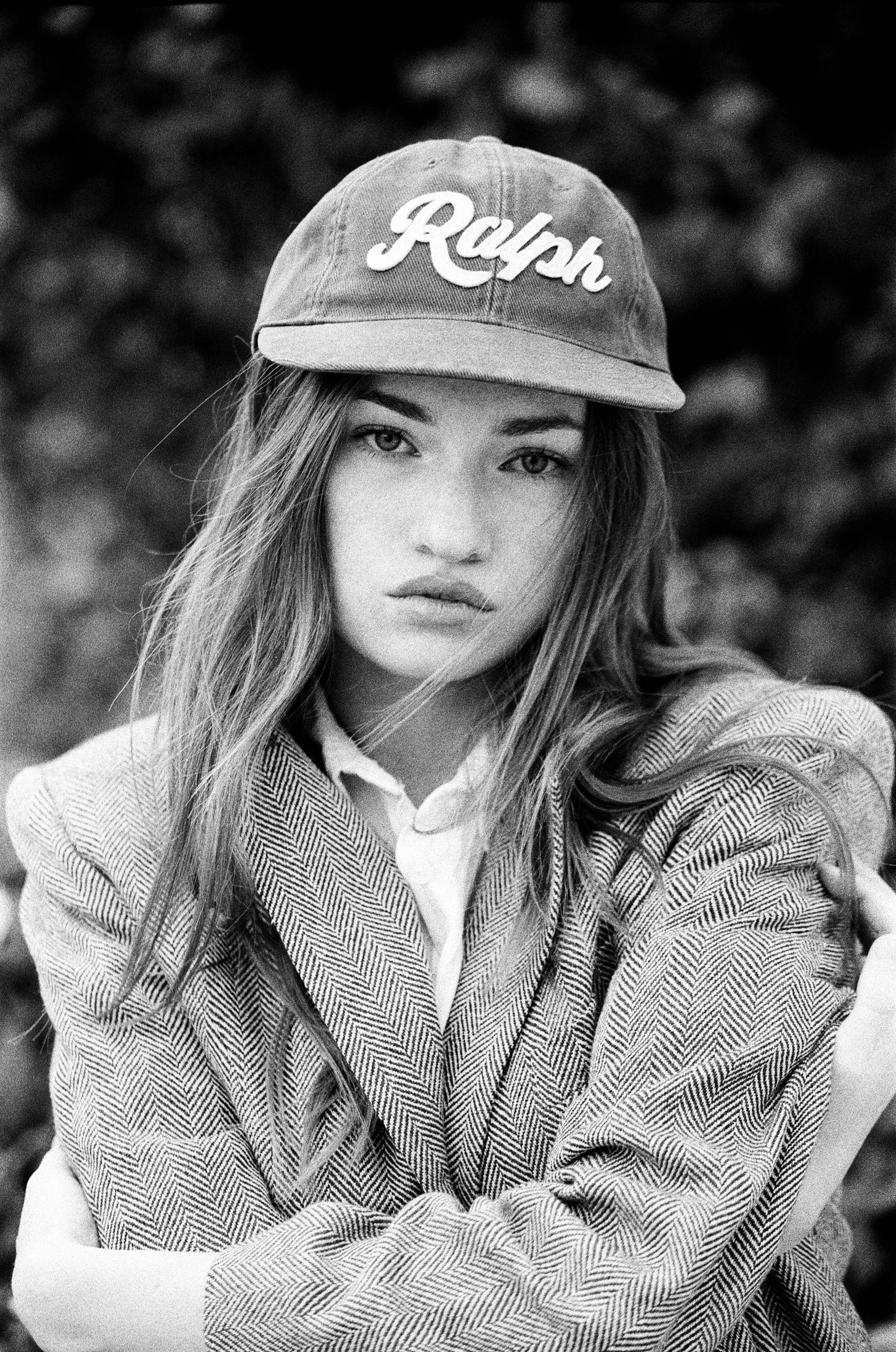 Black and white photo of a young woman wearing a baseball cap with the word 'Ralph' on it, a collared shirt, and a jacket, with long hair and striking eyes, outdoors.