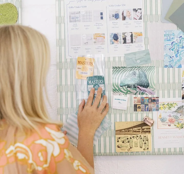 A woman with blonde hair in a pink and orange patterned shirt is looking at a bulletin board filled with various images, flyers, and tickets. She is touching a teal-colored ticket.