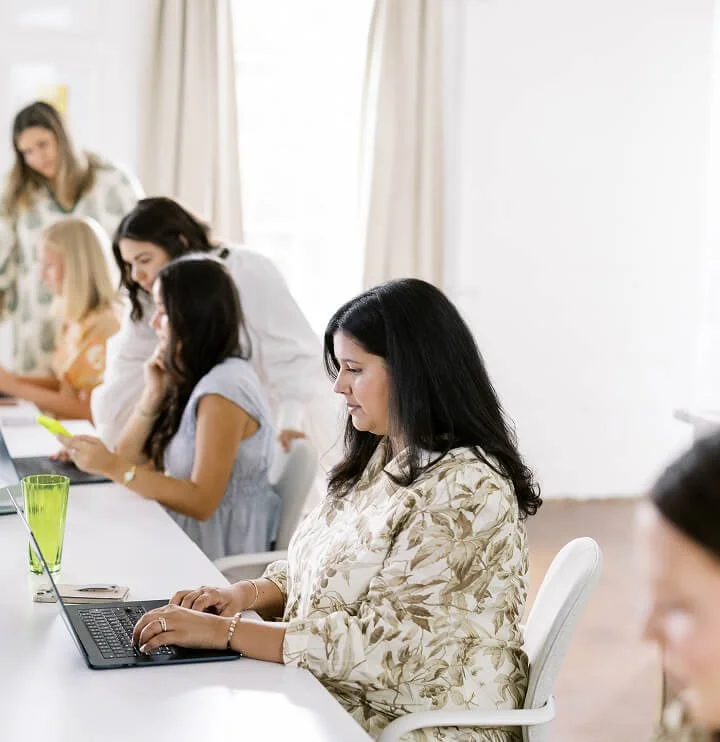 A group of women working at a long white table with laptops and drinks in a bright room.