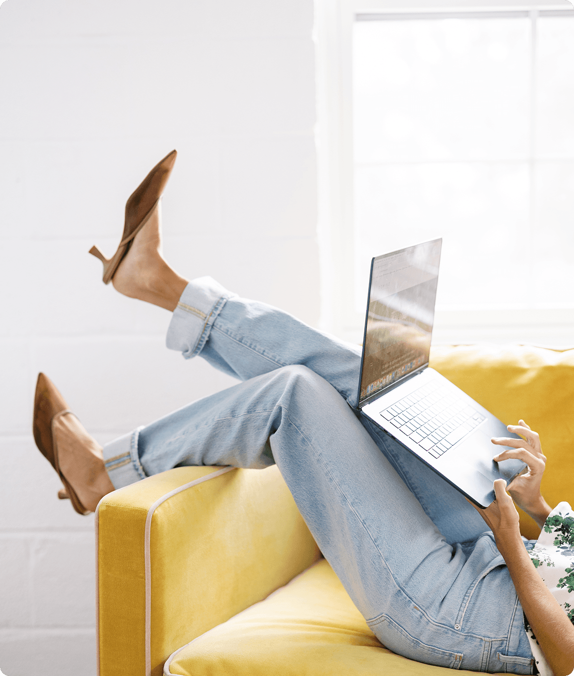 Person lying on a yellow couch with laptop, wearing jeans and brown boots, in a bright room with white walls and large window.