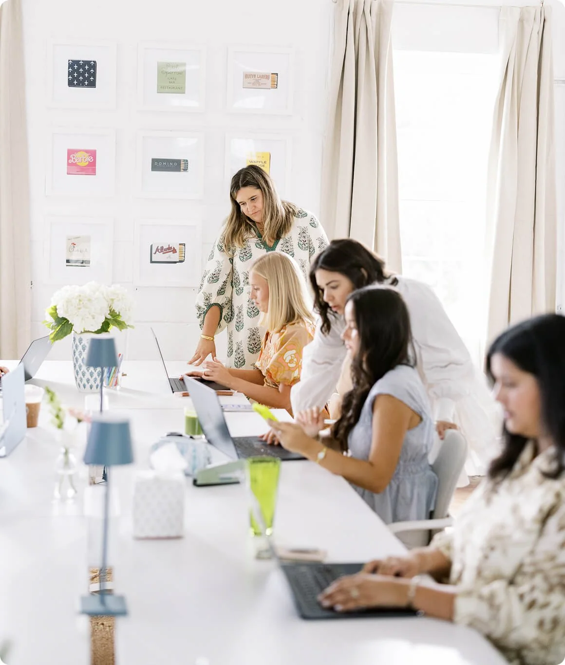 Women working in a bright meeting room with laptops and papers, decorated with flowers and framed pictures on the wall.