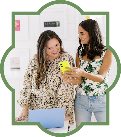 Two women smiling and looking at a smartphone together inside a room with art on the walls and a laptop on the table.