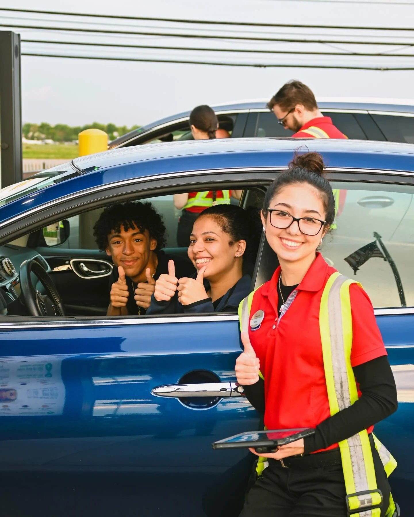 A young woman in a red safety vest standing outside a blue car, giving a thumbs-up, with three people inside the car also giving thumbs-up through the window at a Chick-fil-A drive thru.
