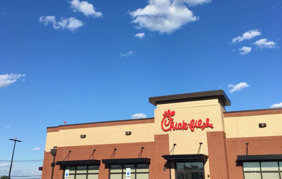 Exterior view of a Chick-fil-A restaurant with a brick and beige facade, large windows, and a sign on the building, under a blue sky with scattered clouds.