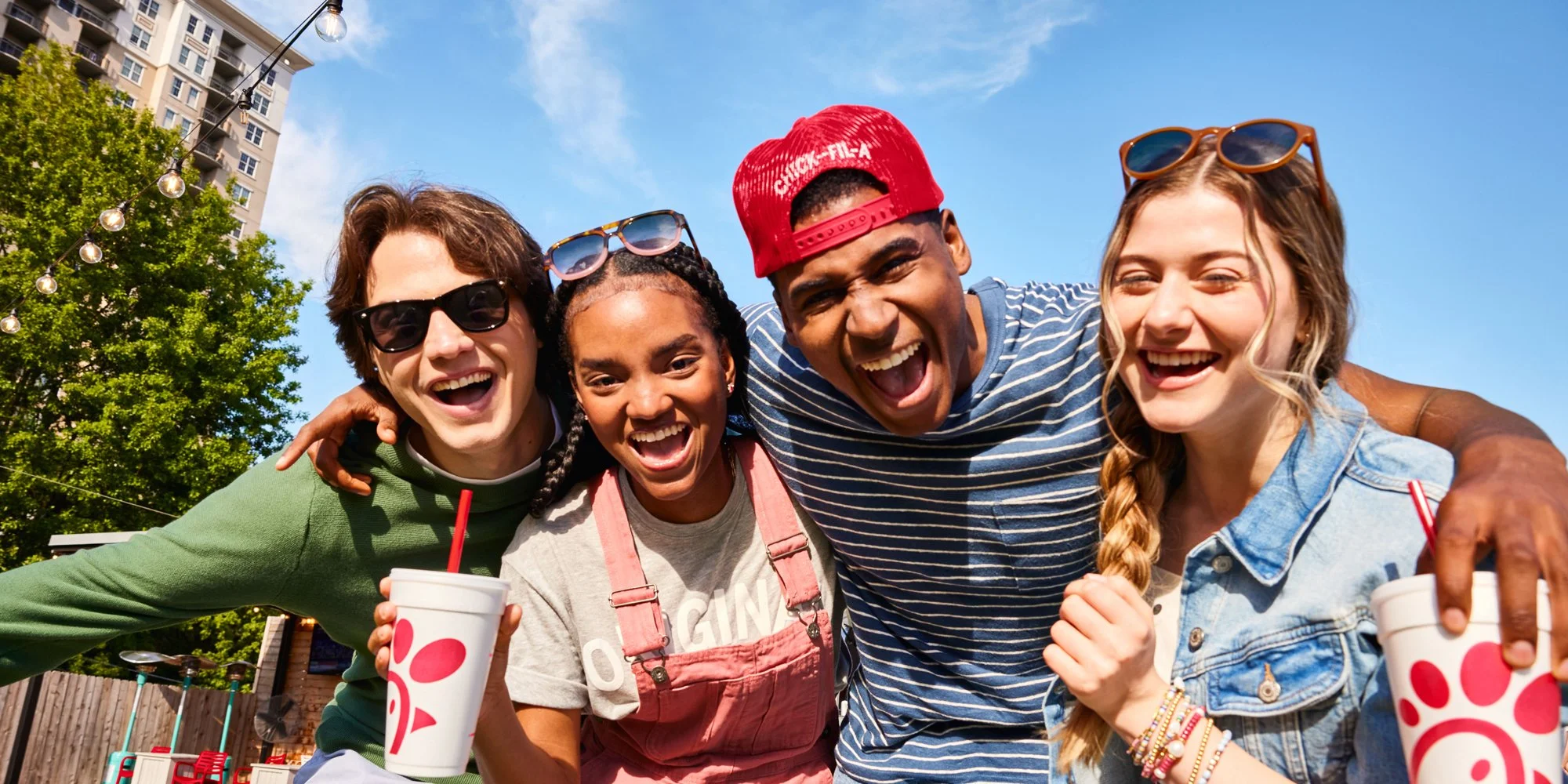 Group of four friends smiling and celebrating outdoors on a sunny day, holding Chick-fil-A cups, with a blue sky, trees, and buildings in the background.