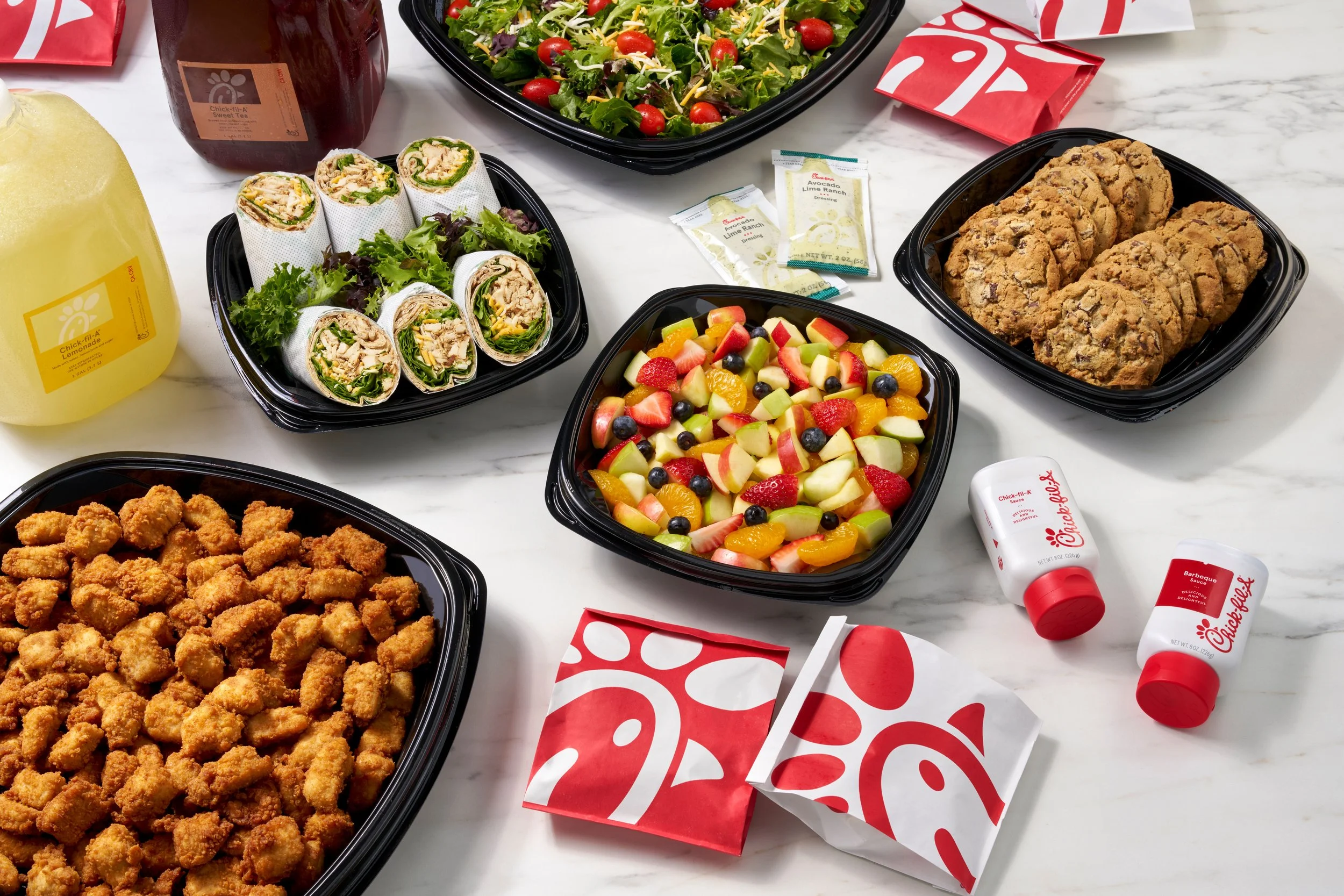 Chick-fil-A food spread on a white table, including fried chicken nuggets, chicken wraps, a mixed fruit salad, a salad with cherry tomatoes, and chocolate chip cookies, with Chick-fil-A sauce and small bottles of dipping sauce.