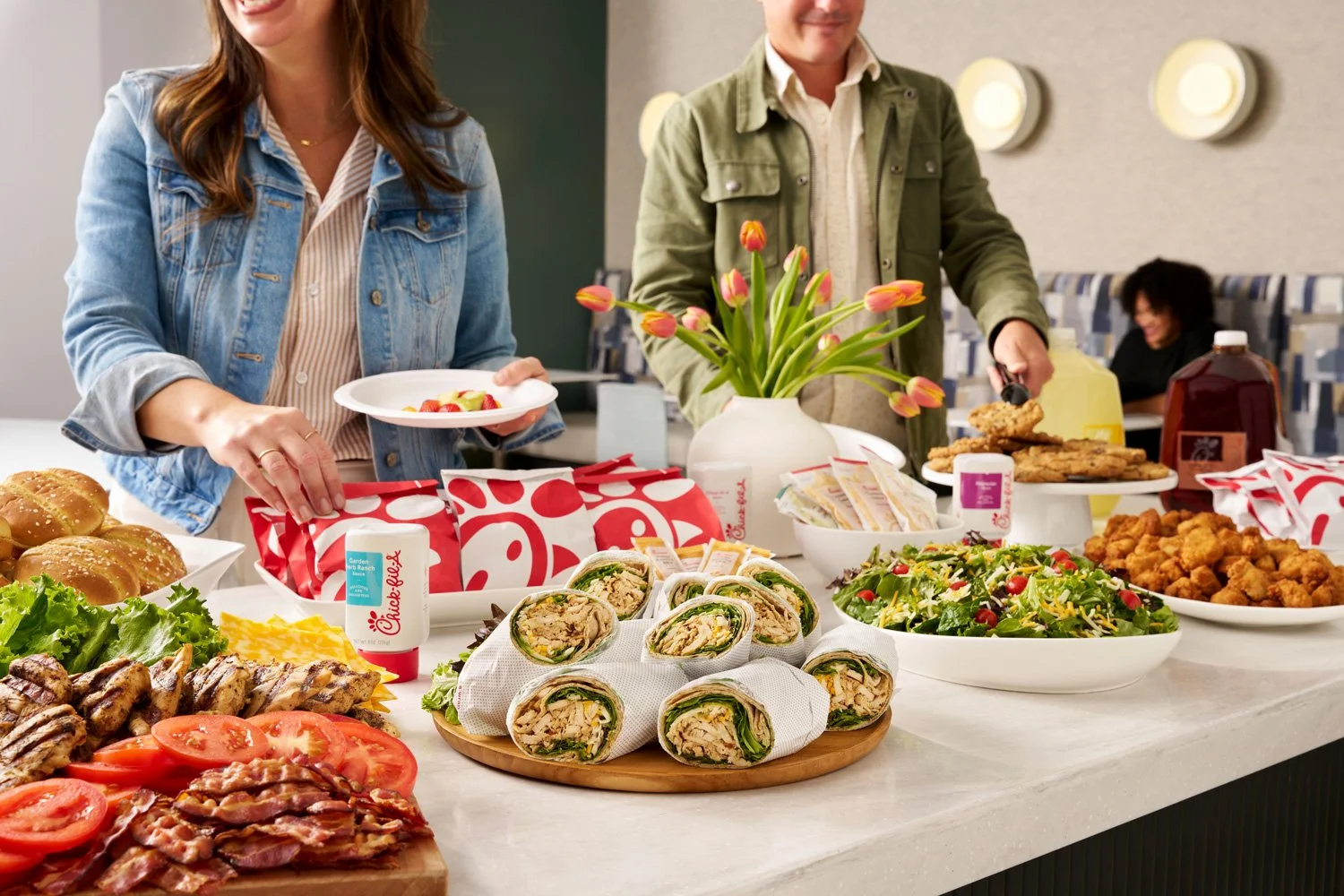 People serving themselves food at a buffet table with various dishes including sandwiches, salads, fried chicken, baked goods, and beverages; a woman is holding a plate of strawberries, and a man is pouring a drink.
